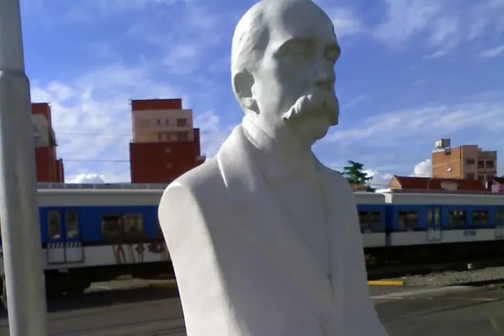 En primer plano, un busto de un hombre con barba y bigote, con un tren detrás