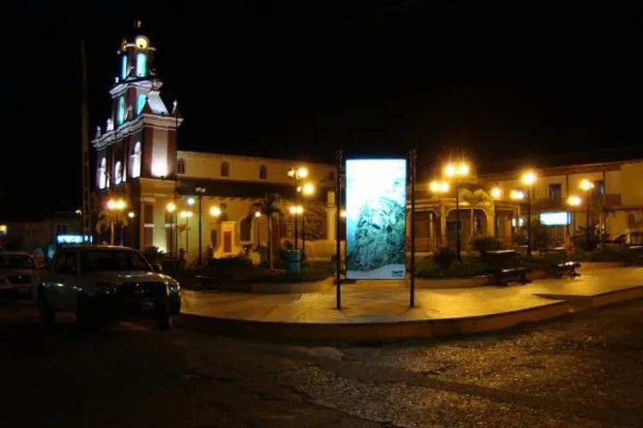 En primer plano, una plaza con una iglesia en la esquina, iluminada por farolas de luz amarilla