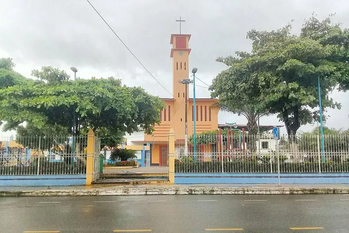 En primer plano, una iglesia con una torre y un reloj, rodeada de árboles y una valla metálica