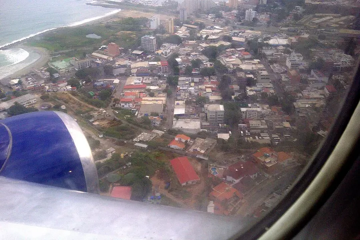 En primer plano, un motor de avión, con una vista aérea de una ciudad costera con edificios y casas