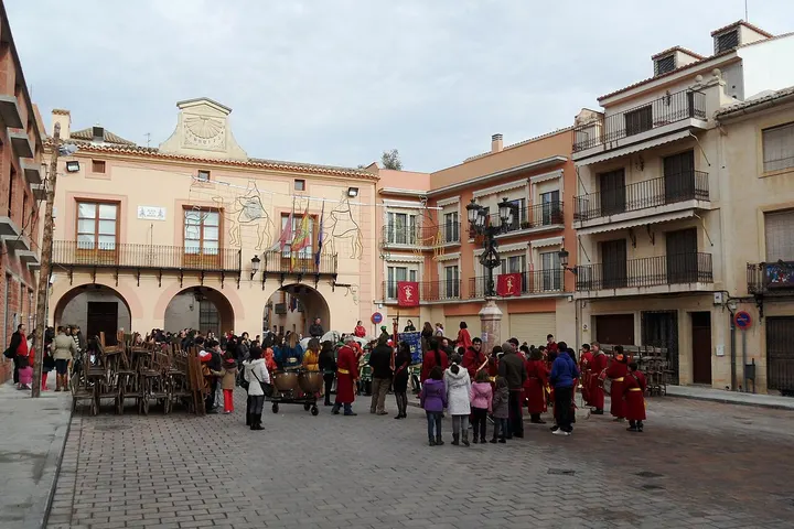 En primer plano, una procesión con carros de madera y personas vestidas de rojo y blanco