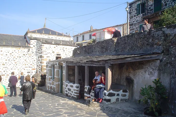 En primer plano, un hombre con una guitarra eléctrica en la calle de una ciudad con casas de piedra