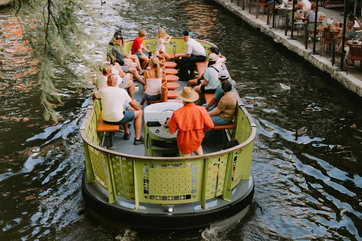 Un grupo de personas en un barco de turismo, navegando por un canal, en un lugar con árboles y restaurantes