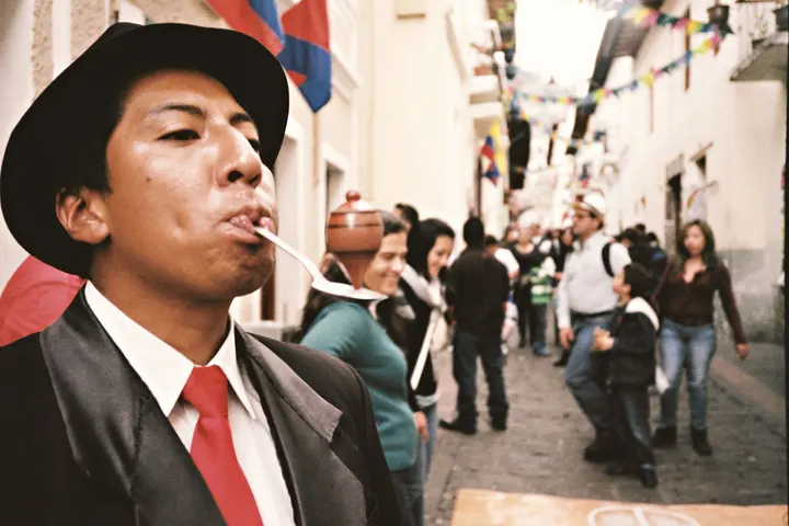 Un hombre de sombrero y traje negro fuma un cigarrillo en una calle de una ciudad, rodeado de gente