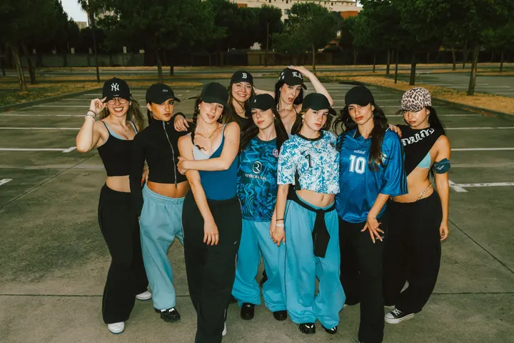 Un grupo de nueve mujeres jóvenes, vestidas con ropa deportiva y gorras, posan en un aparcamiento