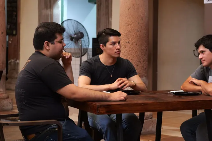 Tres hombres con ropa oscura se sientan a una mesa en un lugar con columnas y un ventilador