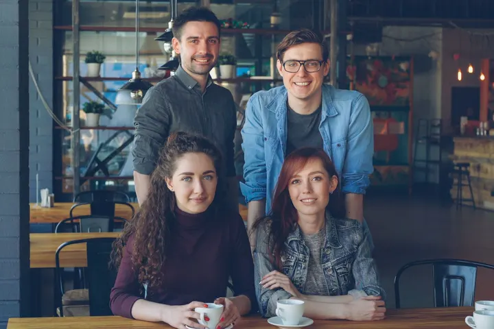 Cuatro personas se encuentran en un café, sentadas en una mesa, mientras uno de ellos se encuentra detrás de ellas