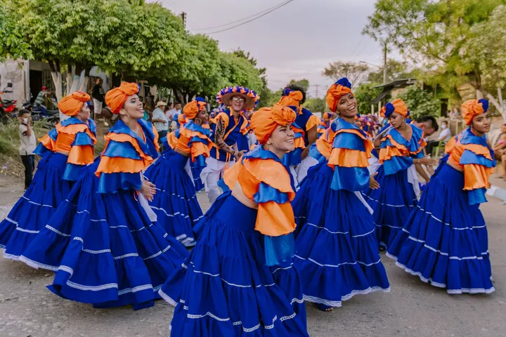 Un grupo de mujeres en trajes tradicionales de colores azul y naranja bailan en una calle con árboles